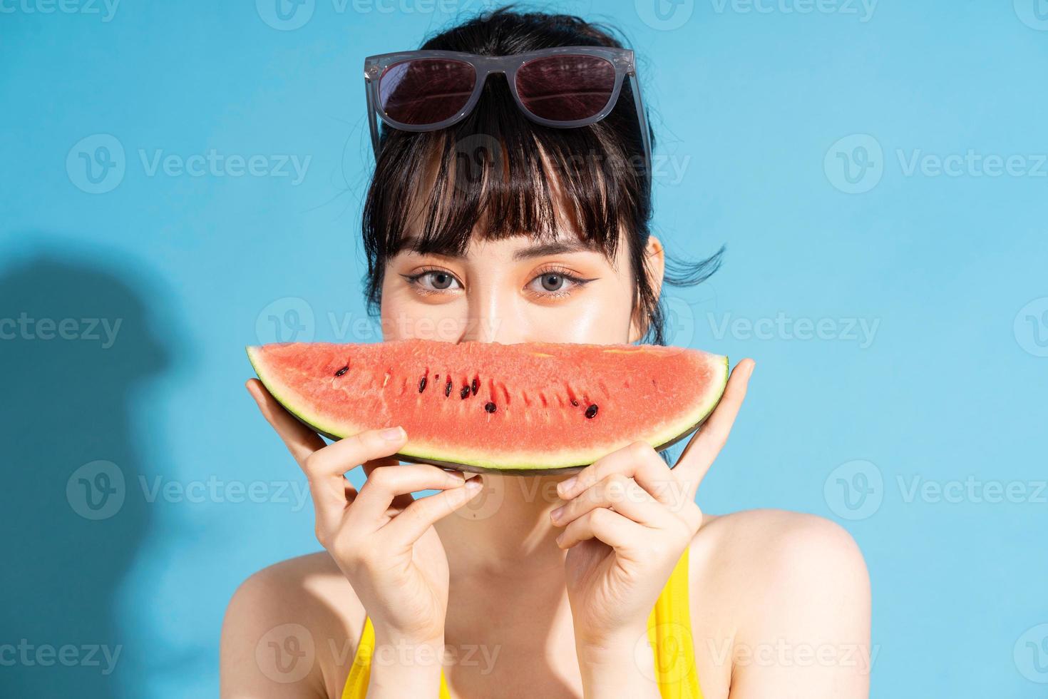 linda mulher asiática vestindo macacão amarelo sobre fundo azul e comendo frutas tropicais, conceito de verão foto