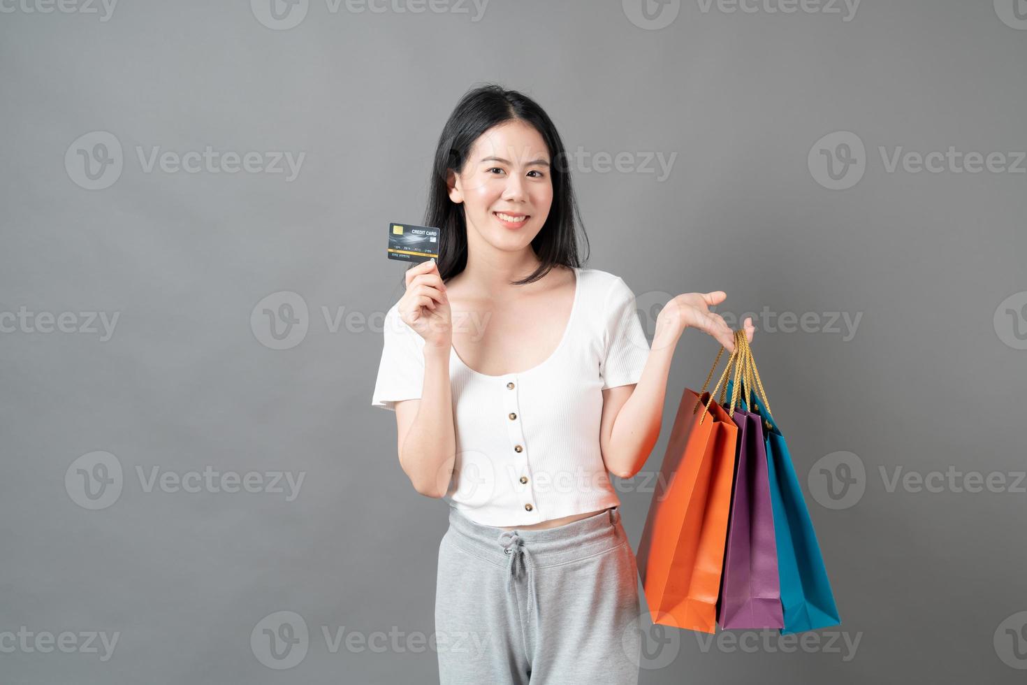 jovem mulher asiática segurando uma sacola de compras e um cartão de crédito em fundo cinza foto