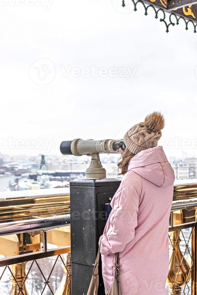 uma jovem olha através de binóculos operados por moedas no deck de observação com vista para a cidade de uma altura ao pôr do sol. inverno, queda de neve foto