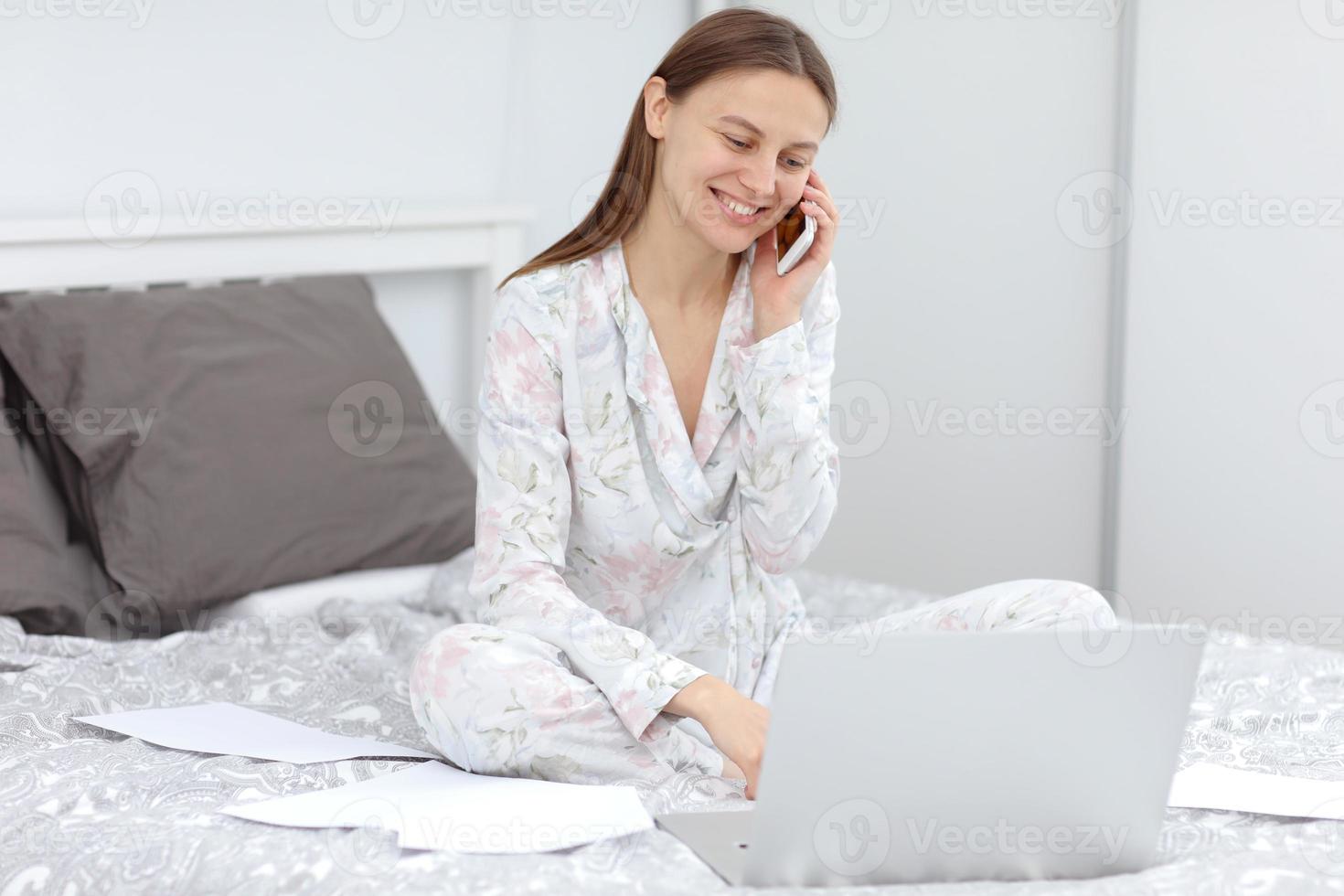 mulher jovem e atraente trabalhando em casa - empreendedora sentada na cama com o computador laptop, papelada e falando ao telefone celular do conforto de casa. quarentena. coronavírus foto