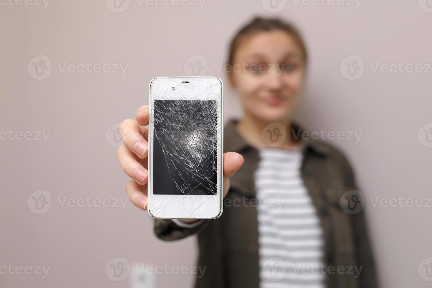 jovem frustrada segurando o celular com o vidro da tela quebrada. a tela do telefone precisa ser consertada. isolado em fundo cinza. foco seletivo no smartphone foto