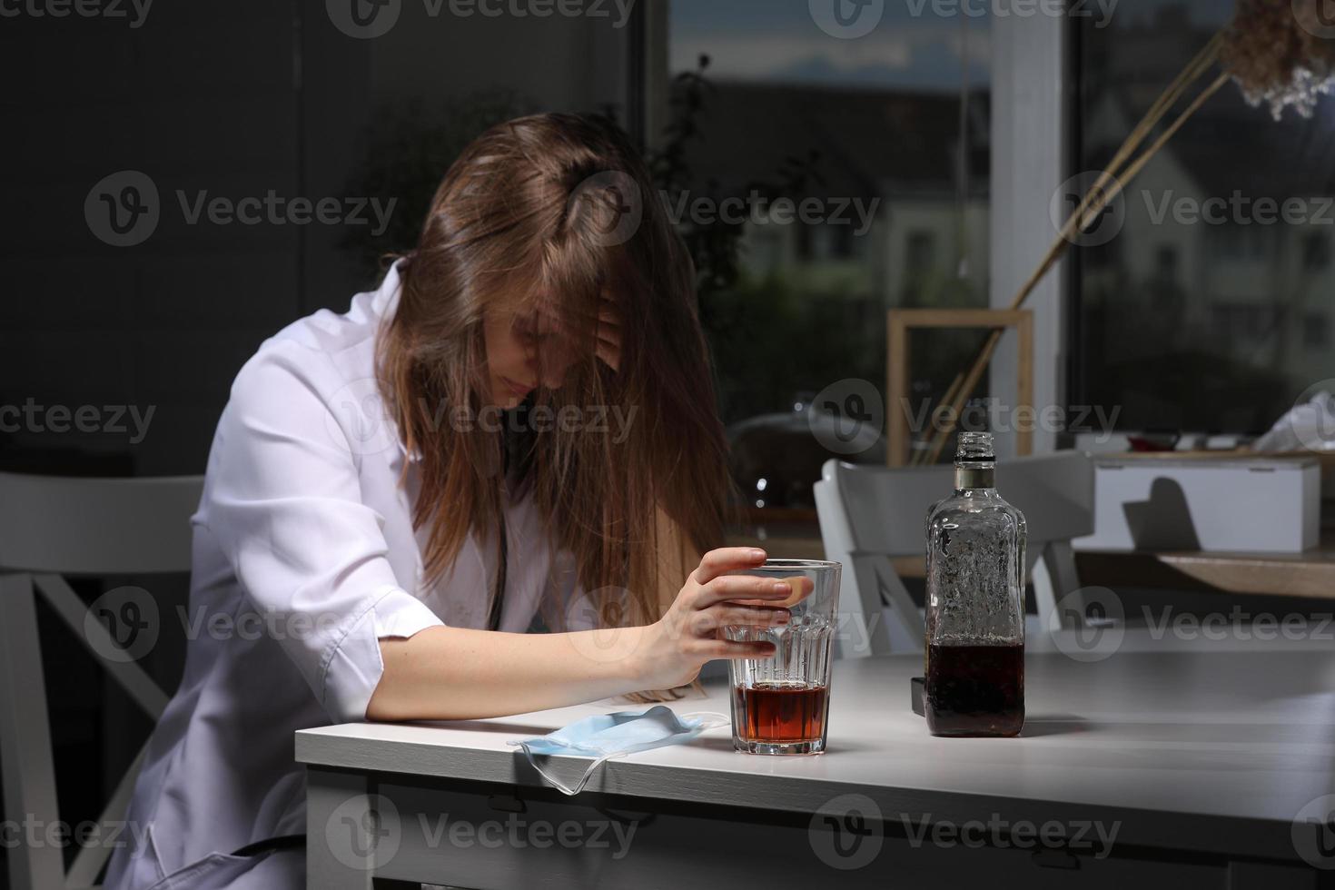 médica sentada à mesa e segura o copo com uísque ou conhaque em casa depois do trabalho duro, mulher deprimida bebendo álcool forte, sofrendo de problemas de coronavírus. foto