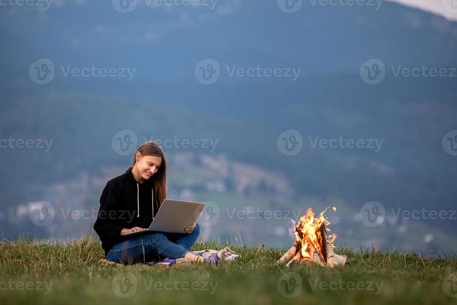 jovem freelancer com laptop nas montanhas à noite. turista garota sentada perto da fogueira foto