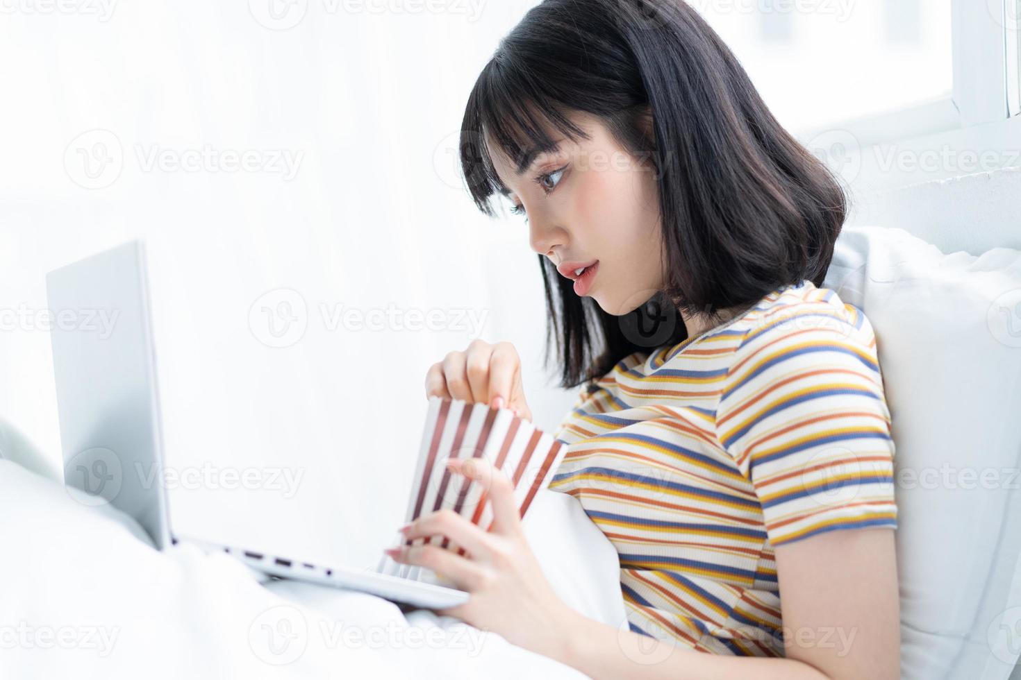 jovem asiática usando laptop para assistir programas de TV, comendo pipoca em casa foto