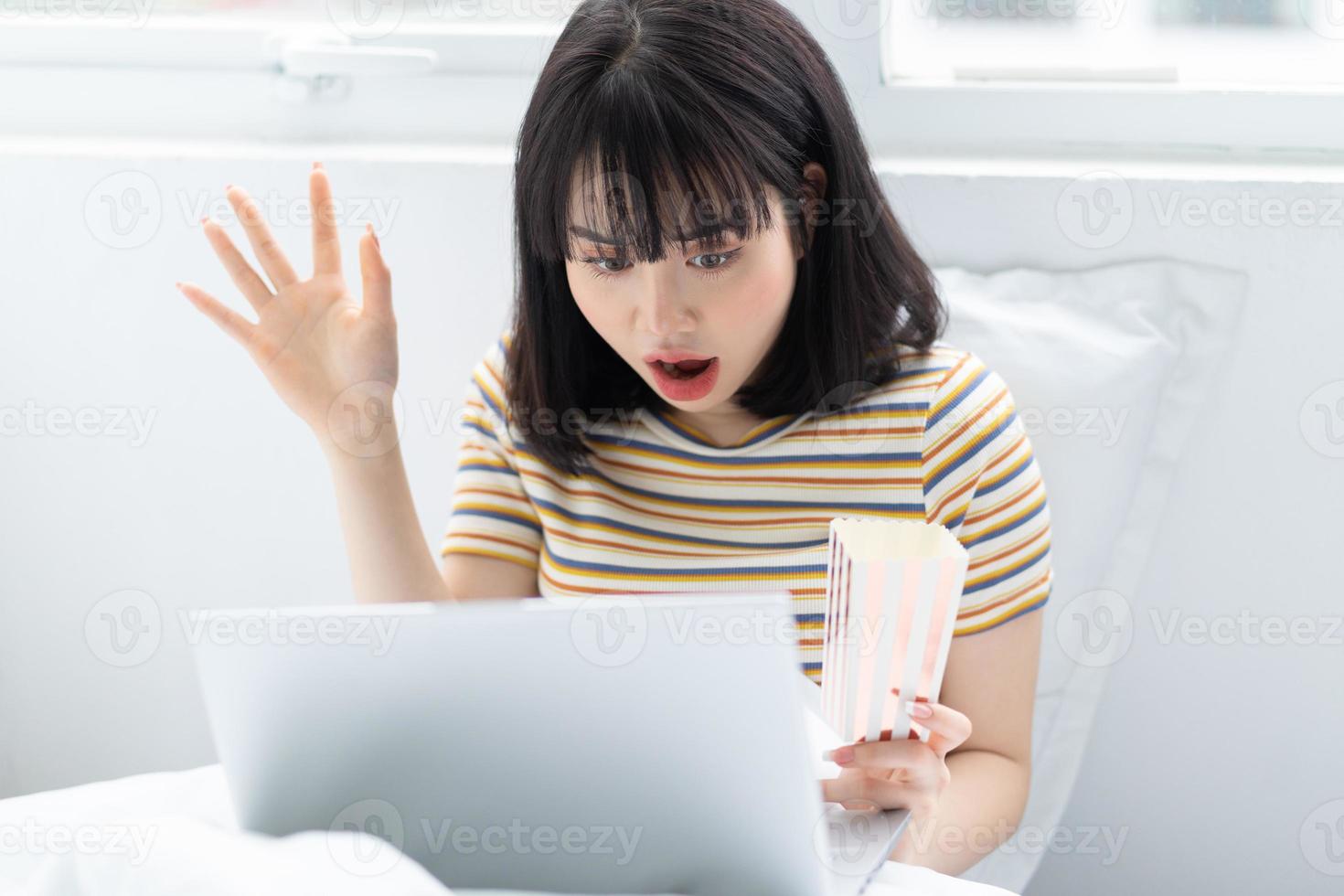 jovem asiática usando laptop para assistir programas de TV, comendo pipoca em casa foto