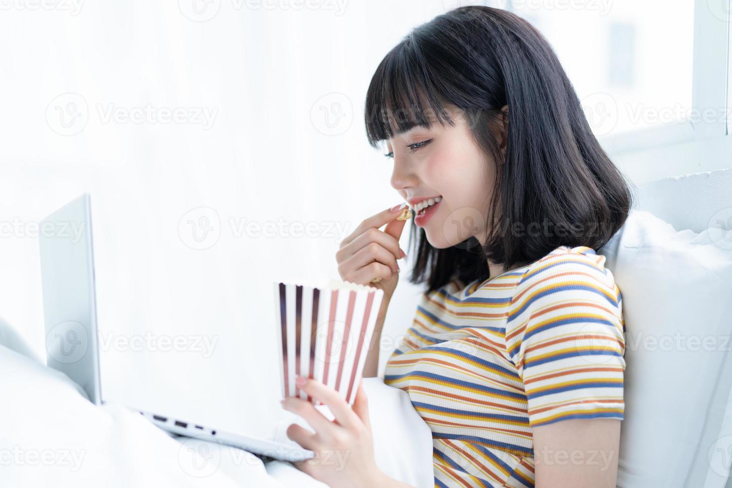 jovem asiática usando laptop para assistir programas de TV, comendo pipoca em casa foto