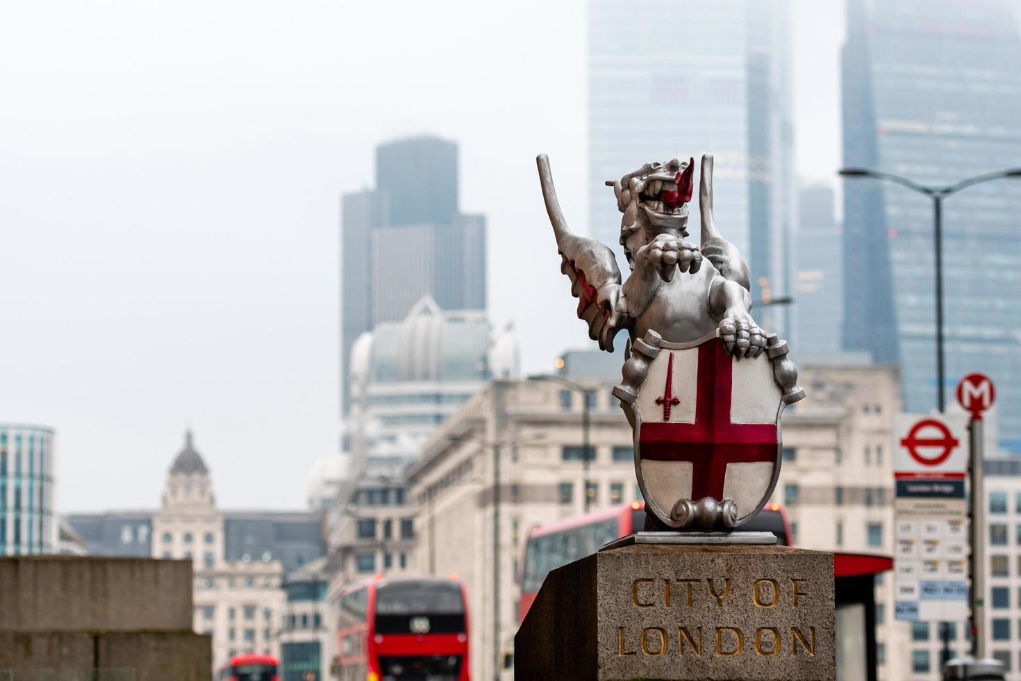 uma escultura de dragão com o brasão de armas da cidade de londres na ponte de londres. arranha-céus borrados e ônibus vermelhos de dois andares ao fundo. foto