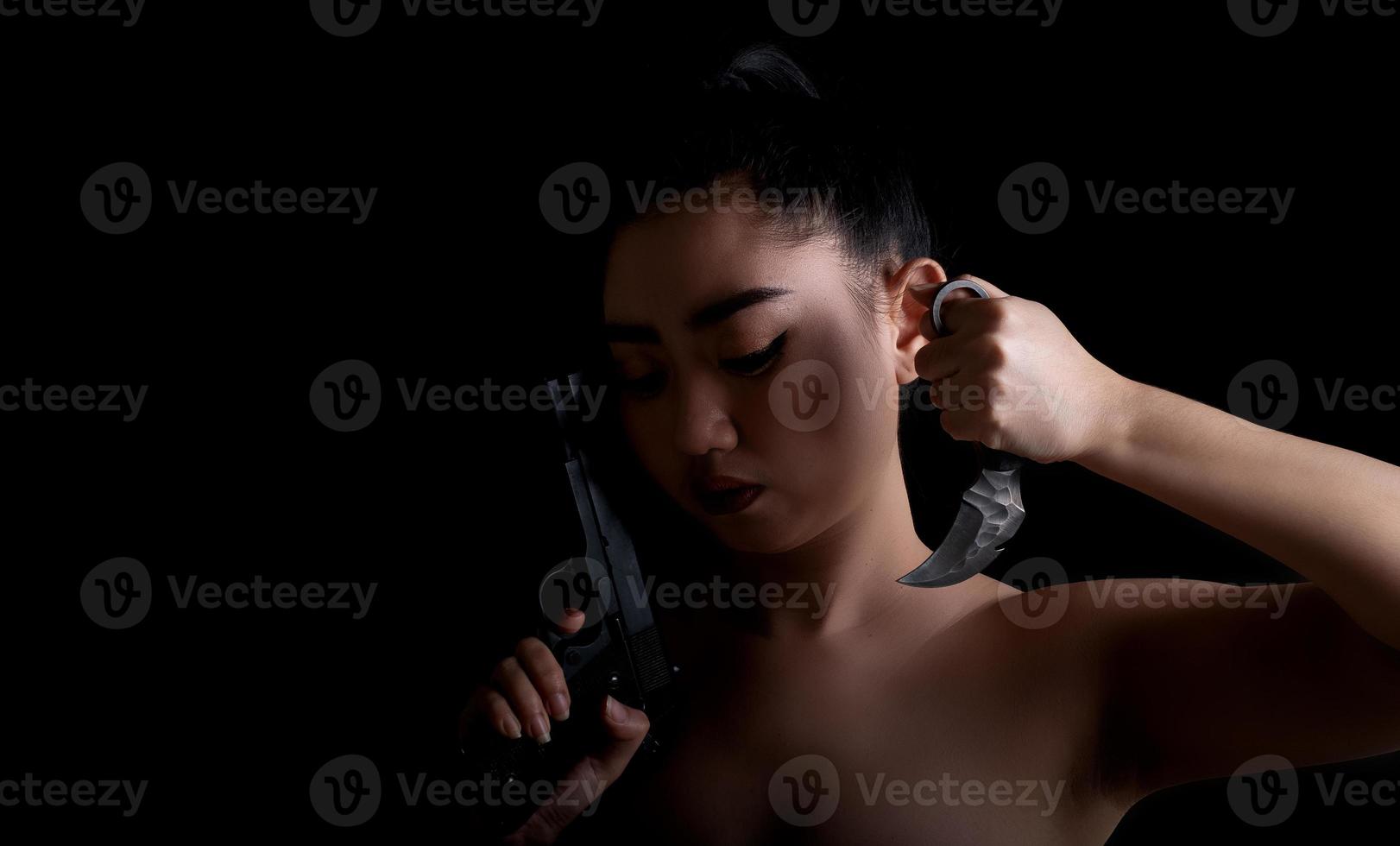 Ásia mulher com uma mão segurando uma arma e uma faca de karambit no fundo preto foto