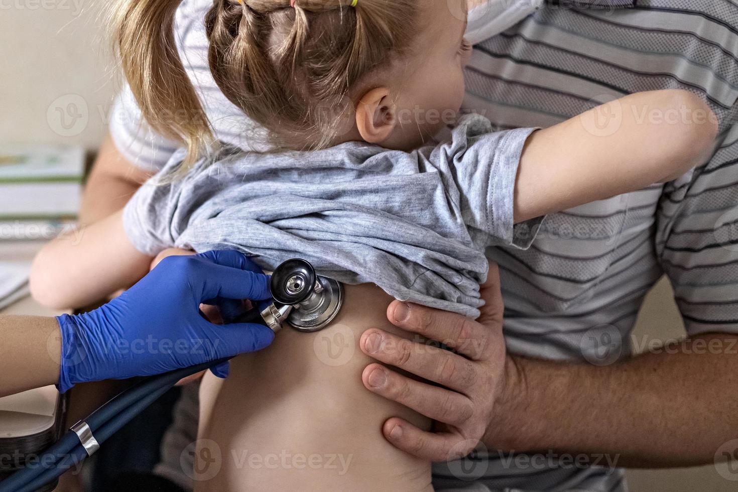 menina nos braços de seu pai no consultório médico na clínica. o médico examina a criança, ouve os pulmões com um estetoscópio. tratamento e prevenção de infecções respiratórias. foto