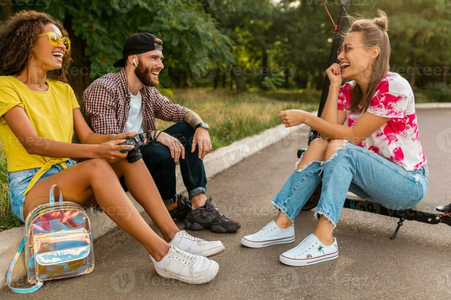 feliz jovem companhia do sorridente amigos sentado dentro parque em Relva com elétrico pontapé lambreta foto