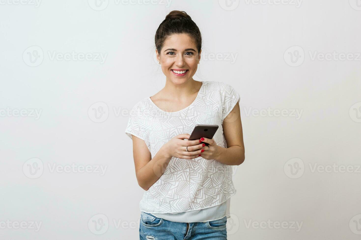 jovem bonita mulher sorridente dentro branco camiseta em branco fundo, segurando e usando Smartphone, Móvel dispositivo, isolado foto