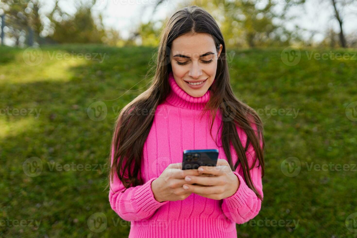 jovem sorridente mulher dentro Rosa suéter caminhando dentro verde parque usando telefone foto