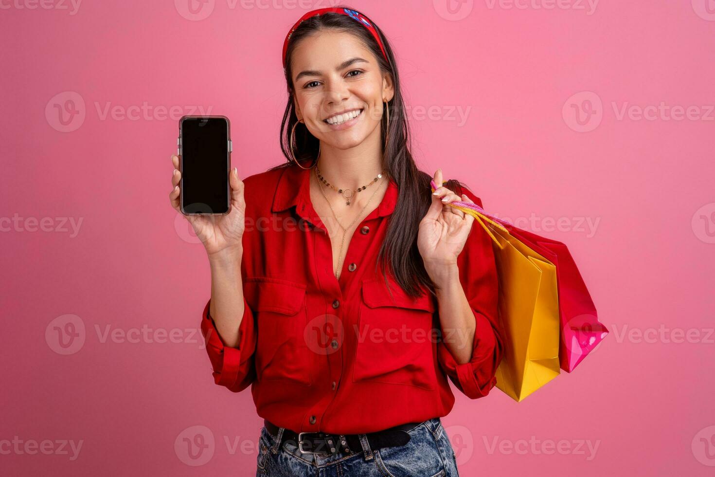 hispânico lindo mulher dentro vermelho camisa sorridente segurando segurando compras bolsas e Smartphone foto