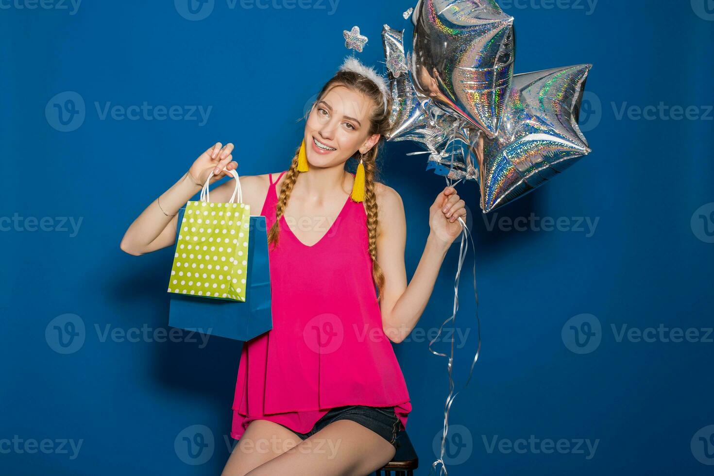 legal jovem hipster lindo mulher, colorida roupa, tendência moda estilo, azul fundo, segurando compras papel bolsas, prata Estrela balões, presentes, sorridente, feliz, a comemorar aniversário foto