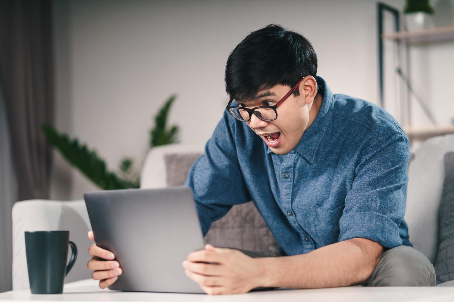 homem asiático chocado em óculos, olhando para o laptop sentado no sofá da sala de estar. foto