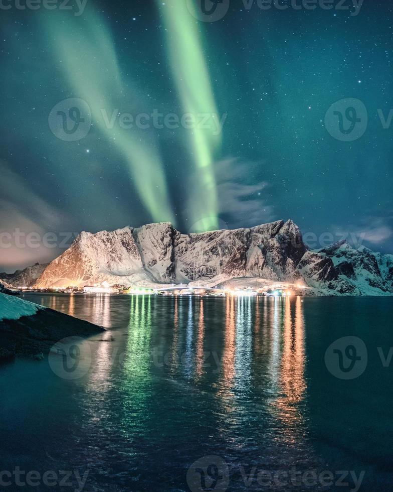 Aurora boreal, aurora boreal sobre uma montanha nevada com uma vila brilhante em Hamnoy nas ilhas Lofoten foto