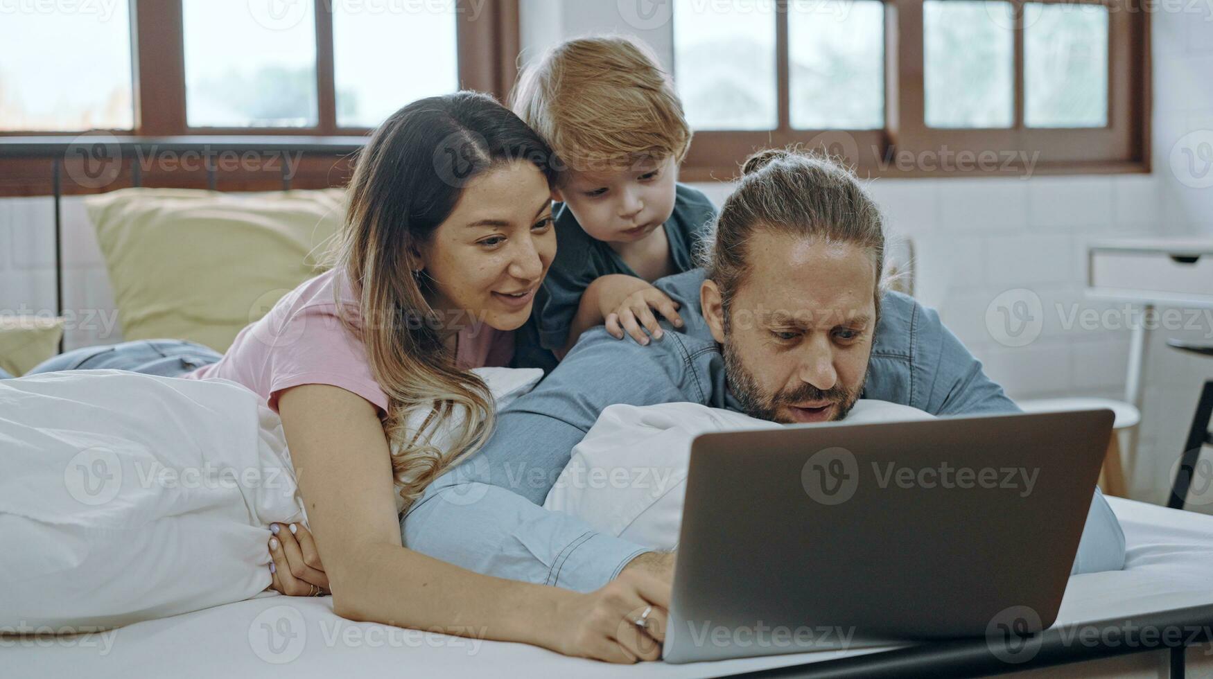 caucasiano família do três usando computador portátil enquanto deitado em cama junto, navegando Internet ou assistindo filme. foto