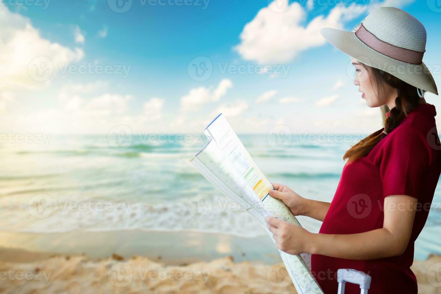 ásia mulher grávida dentro vermelho vestir carregando vermelho bagagem e Veja às a mapa às cedo manhã nascer do sol sobre e onda do a mar em a areia de praia a horizonte plano de fundo, feriado viagem conceito. foto