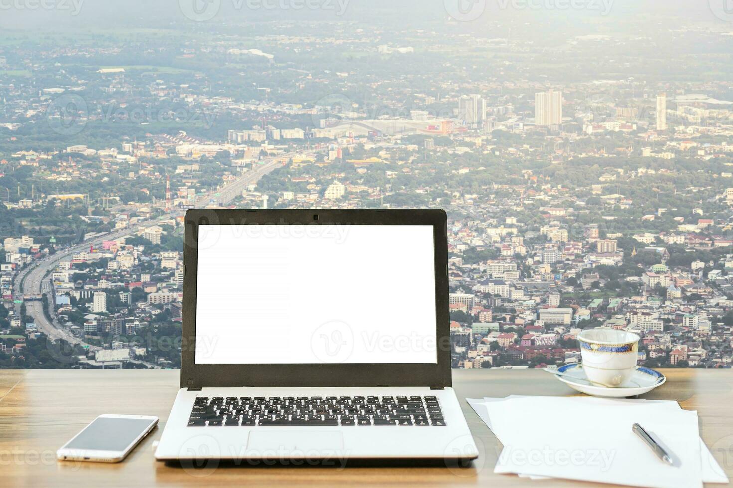 brincar imagem do computador portátil com em branco branco tela, smartphone, café copo em de madeira mesa Visão ao ar livre do escritório construção denso paisagem urbana Alto ângulo Visão fundo. foto