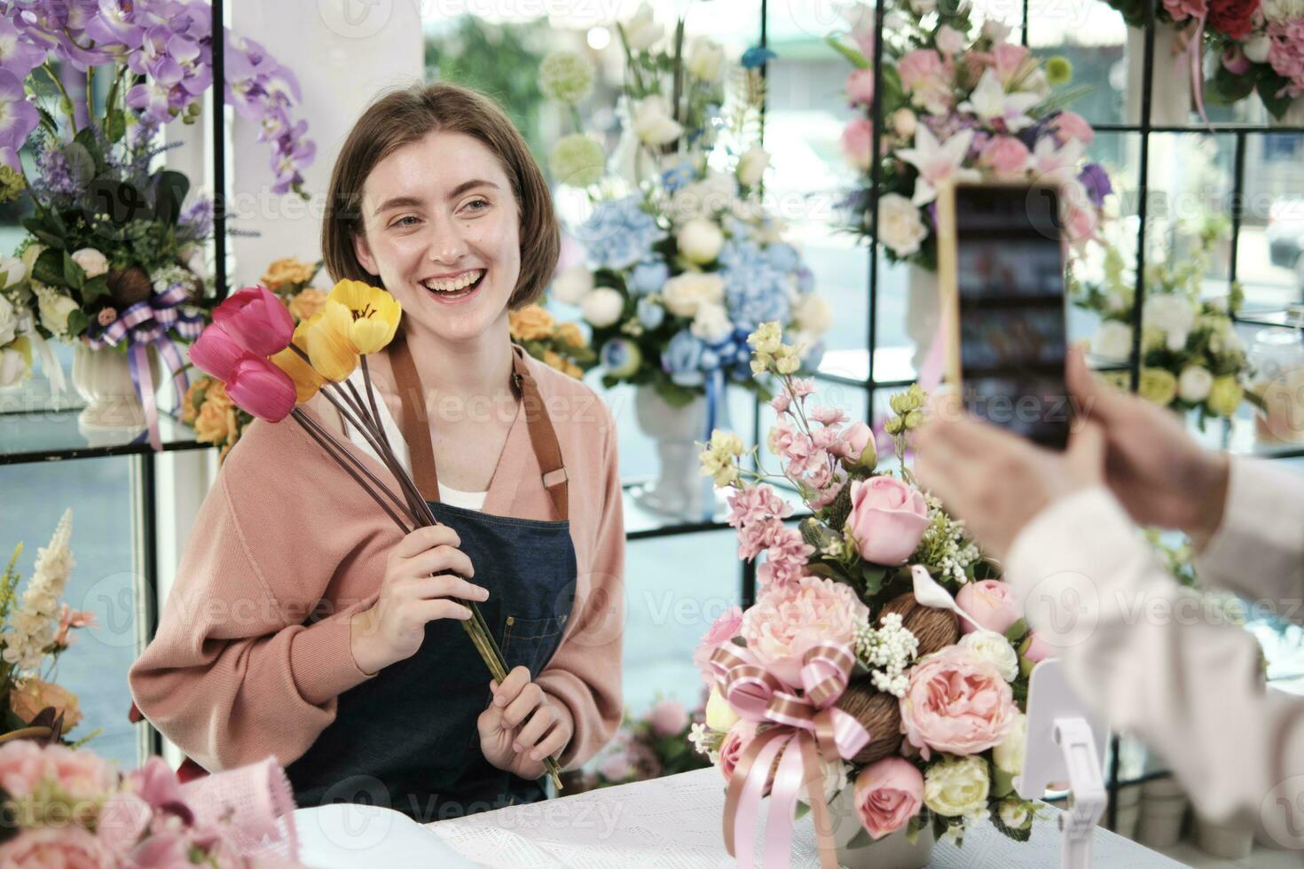 comércio eletrônico negócios, 1 jovem branco fêmea florista demonstra e mostra floral arranjos através da conectados viver transmissão com Smartphone inscrição dentro brilhante flor comprar, lindo flores loja. foto