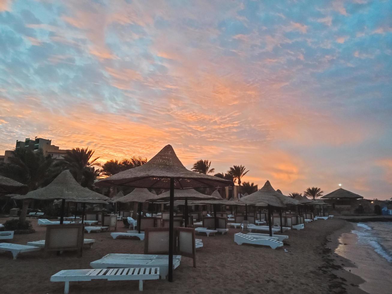 espreguiçadeiras e guarda-sóis de palha e lindo pôr do sol na praia de Hurghada, egito foto