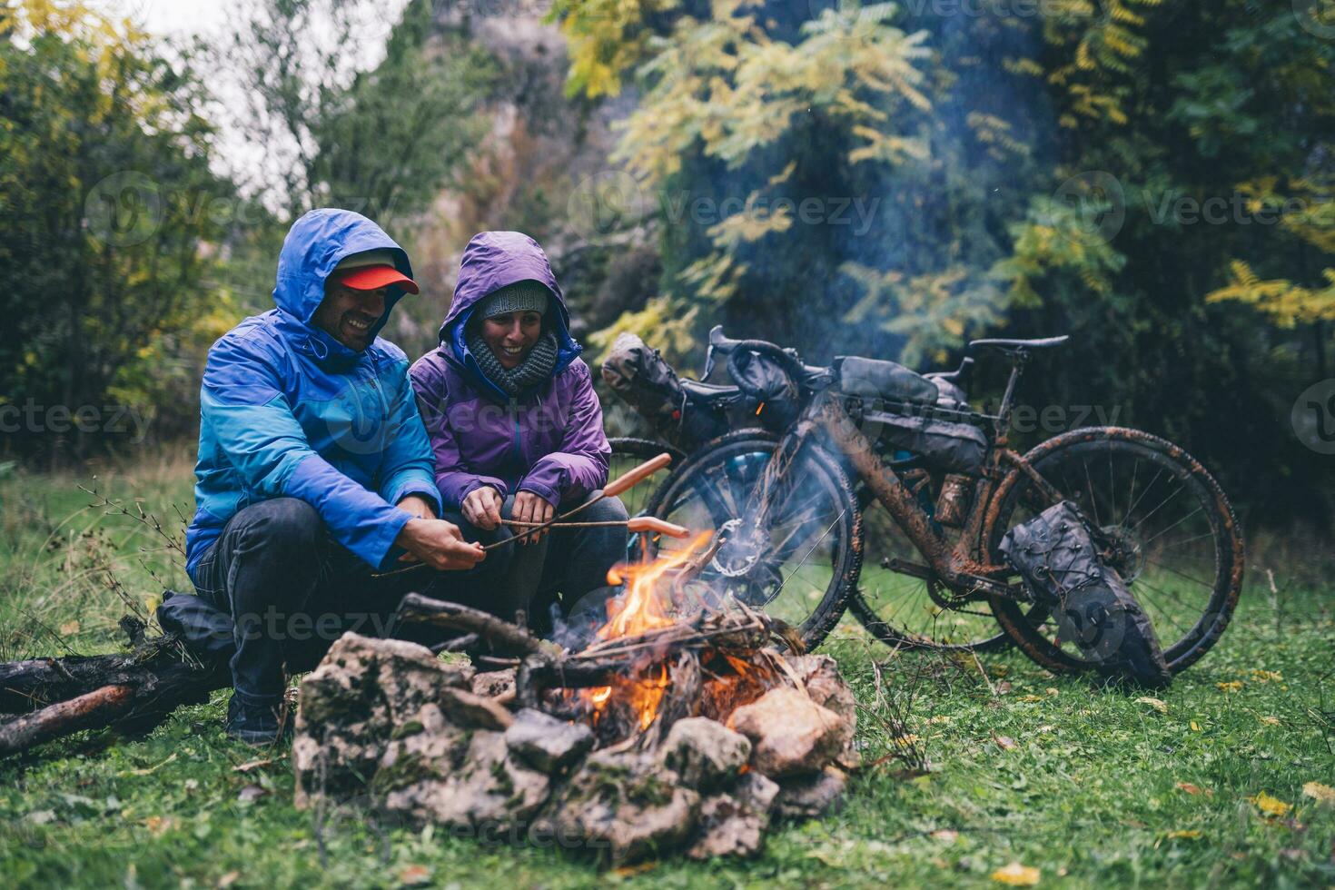 rindo casal com com bmx bicicletas sentado às acampamento fogo grelhar salsichas foto