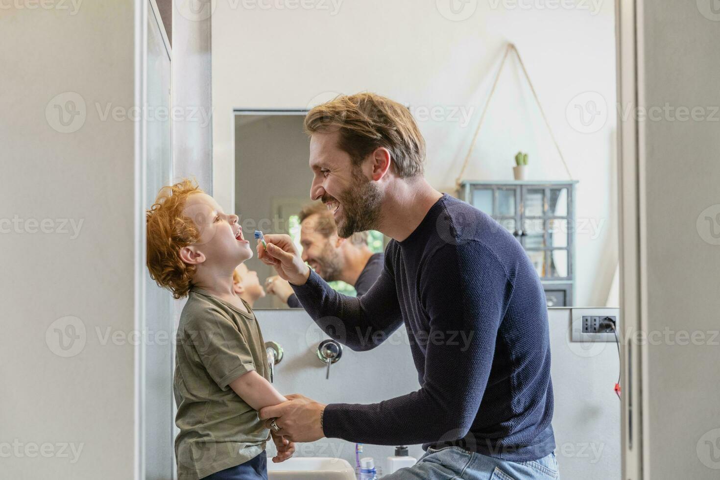 feliz homem escovar filhos dentes com escova de dente dentro banheiro às casa foto