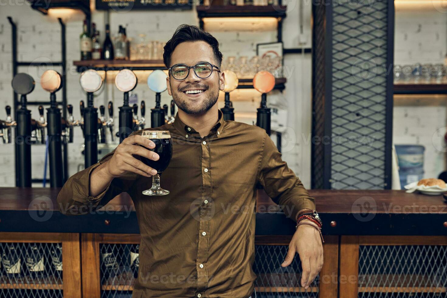 retrato do uma sorridente homem levantando dele Cerveja vidro dentro uma bar foto