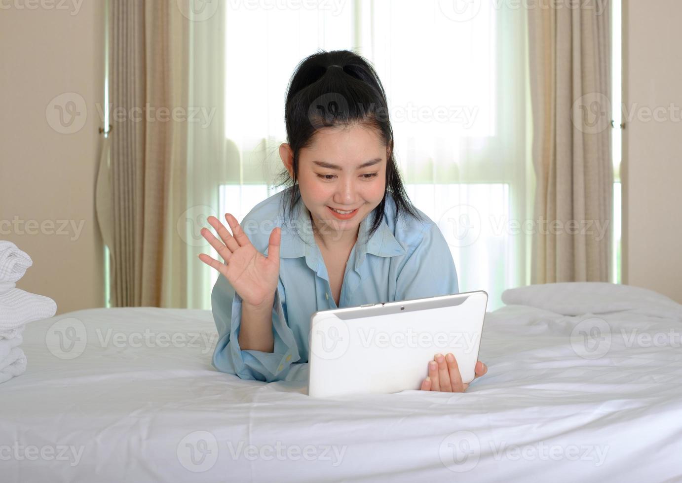 mulheres asiáticas com sorrisos atraentes usam tablet smartphone para videochamadas para dizer oi em uma cama no quarto foto