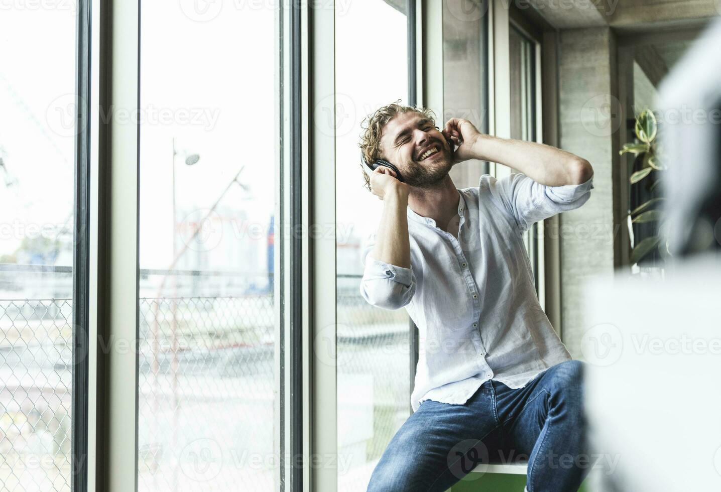 feliz casual jovem homem ouvindo para música com fones de ouvido às a janela foto