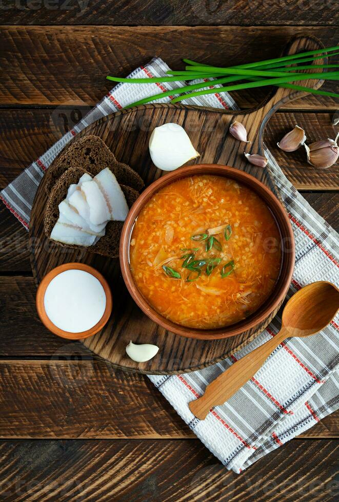 repolho sopa dentro tigela com verde cebola, pão e Salo em de madeira fundo. topo Visão foto