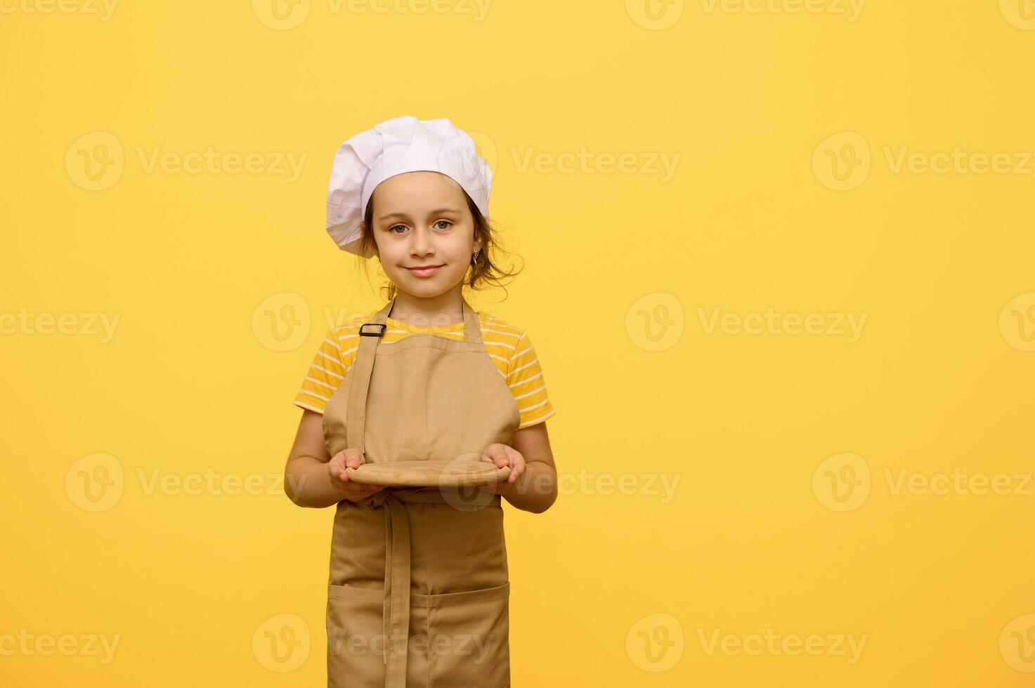 encantador adorável criança menina 6 anos velho, pequeno chefe de cozinha pastelaria, sorrisos fofinho e detém Fora às Câmera uma de madeira borda foto