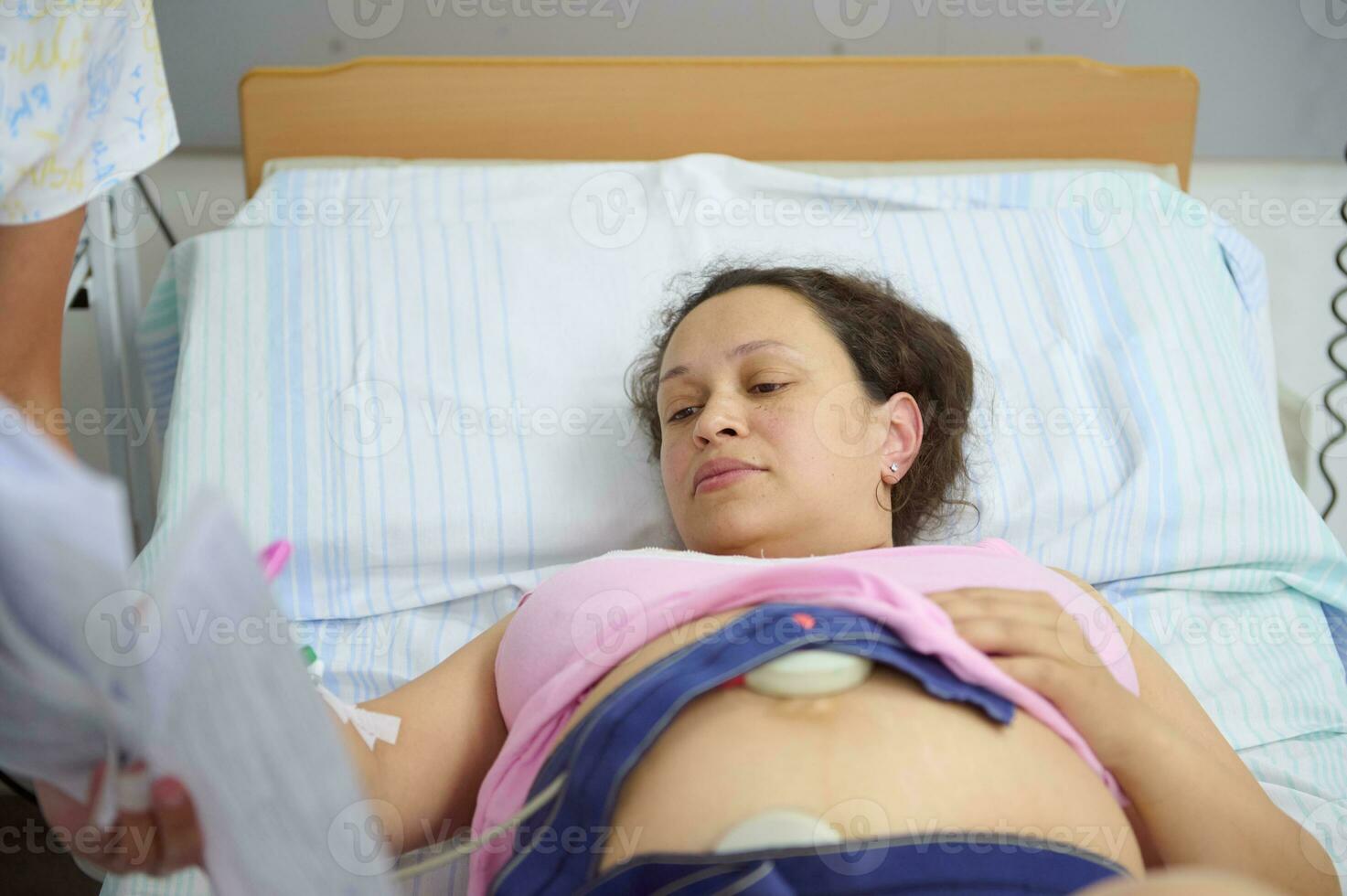 nascimento mãe deitado dentro hospital cama, enquanto uma parteira verificação batimento cardiaco do futuro recém-nascido criança, antes dando nascimento foto
