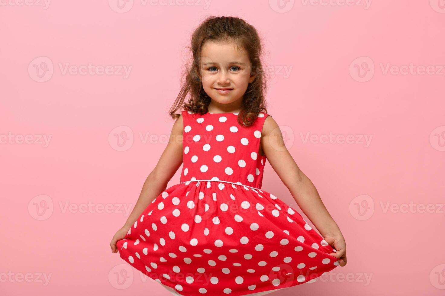 retrato do fofa encantador menina dentro uma Rosa vestir com branco polca pontos posando em rosa fundo fofa sorridente e segurando a inferior do a vestir com dela mãos. lindo criança retrato para publicidade foto