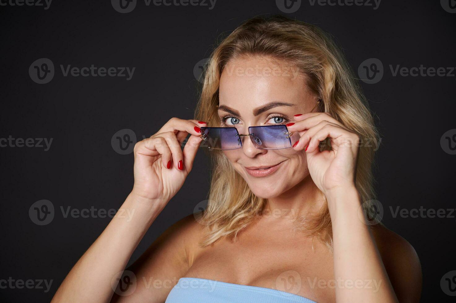 Tiros na Cabeça lindo Loiras europeu mulher com confiança olhando às Câmera através azul oculos de sol, isolado sobre Preto fundo com cópia de espaço para Publicidades foto