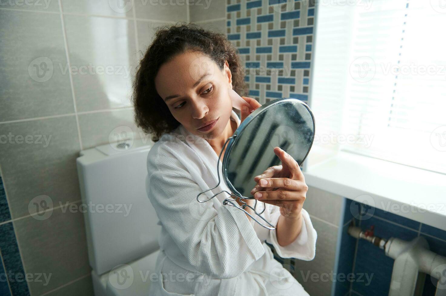 fofa africano americano mulher dentro roupão de banho parece às dela reflexão dentro Cosmético espelho enquanto usando gua sha massagem para linfático drenagem facial massagem foto