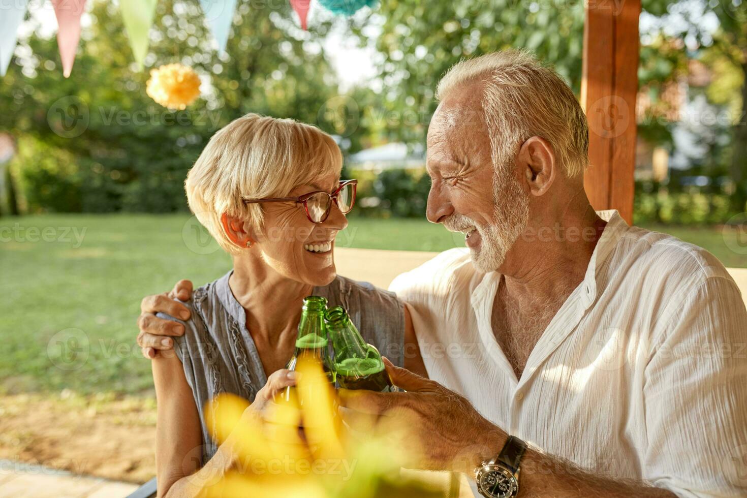 feliz Senior casal tilintar Cerveja garrafas em uma jardim festa foto