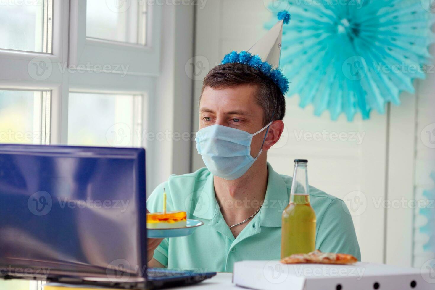 uma homem dentro uma médico mascarar senta dentro frente do uma computador portátil, comemora uma aniversário com amigos sobre a Internet e detém uma vidro do álcool dentro dele mãos. foto