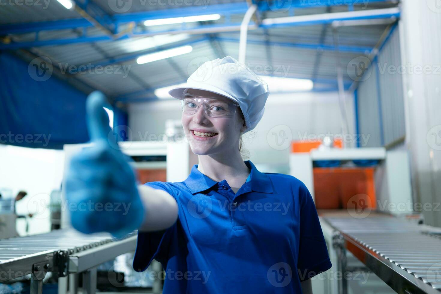 retrato do feliz fêmea fábrica trabalhador mostrando polegares acima gesto dentro bebendo água fábrica foto