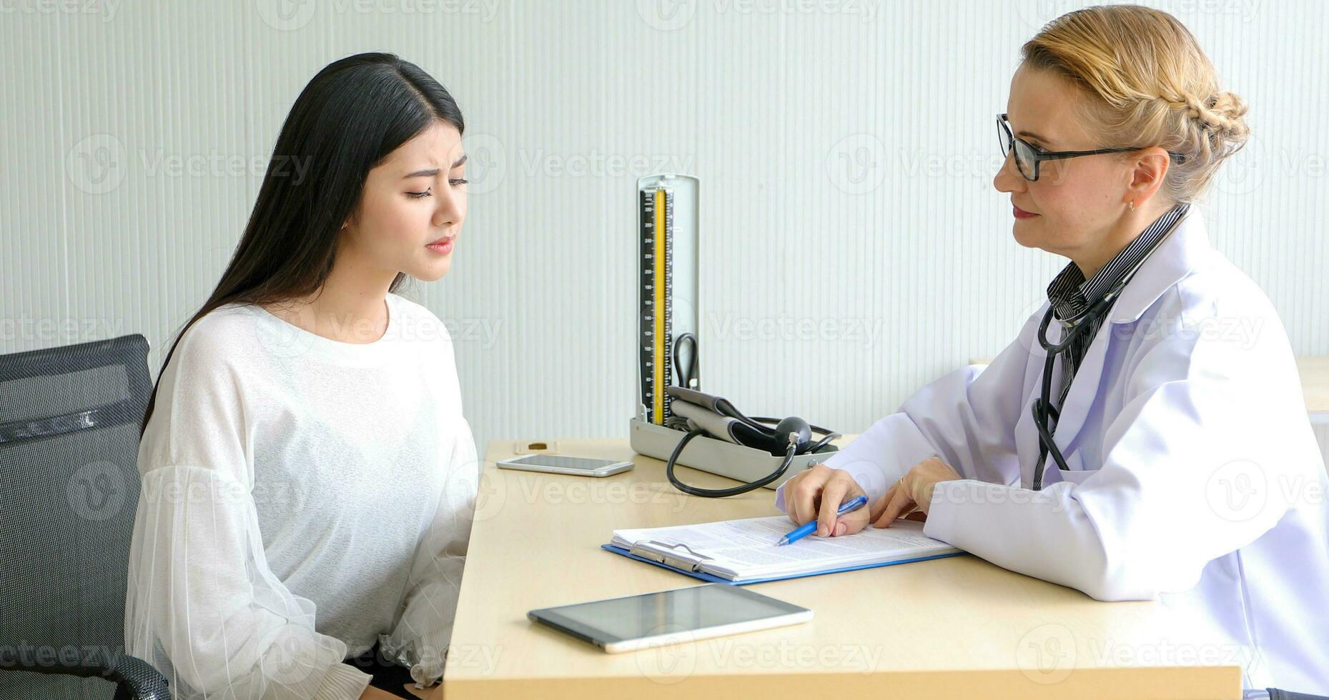 ásia mulher paciente sentado com médico sobre dela doença e mostrando raio X resultados com sangue pressão e coração taxa medição com estetoscópio foto