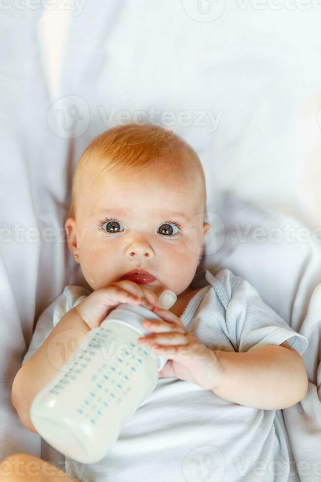 fofa pequeno recém-nascido menina bebendo leite a partir de garrafa e olhando às Câmera em branco fundo. infantil bebê sucção comendo leite nutrição deitado baixa em berço cama às lar. maternidade feliz criança conceito. foto