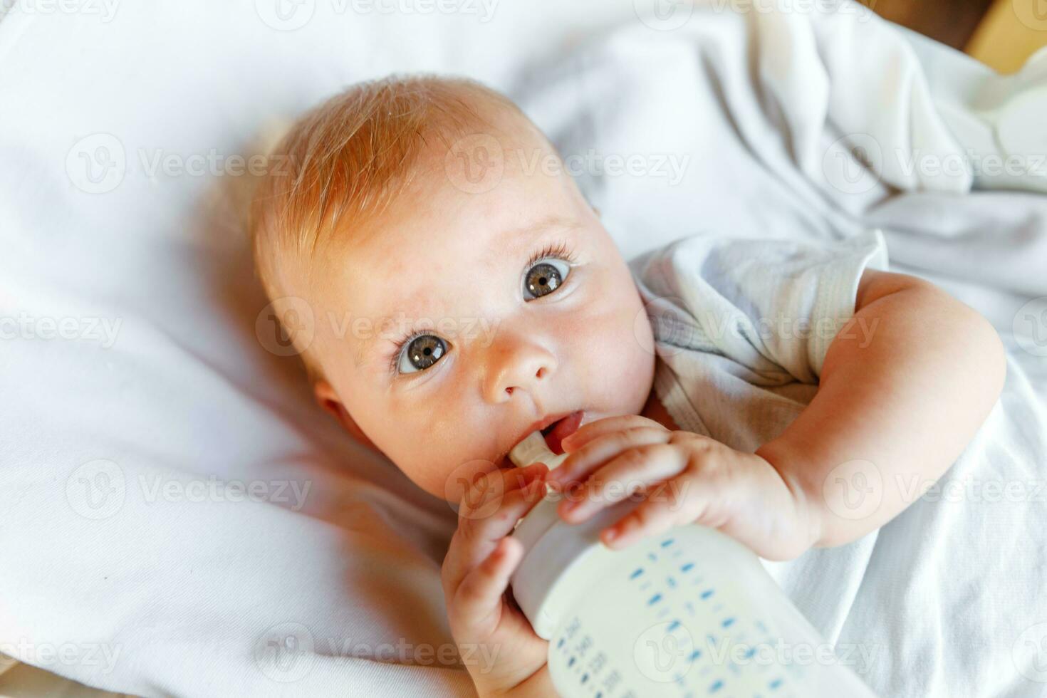 fofa pequeno recém-nascido menina bebendo leite a partir de garrafa e olhando às Câmera em branco fundo. infantil bebê sucção comendo leite nutrição deitado baixa em berço cama às lar. maternidade feliz criança conceito. foto