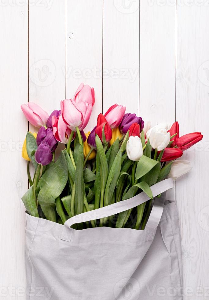 bolsa de tecido cinza cheia de tulipas coloridas em fundo branco de madeira foto