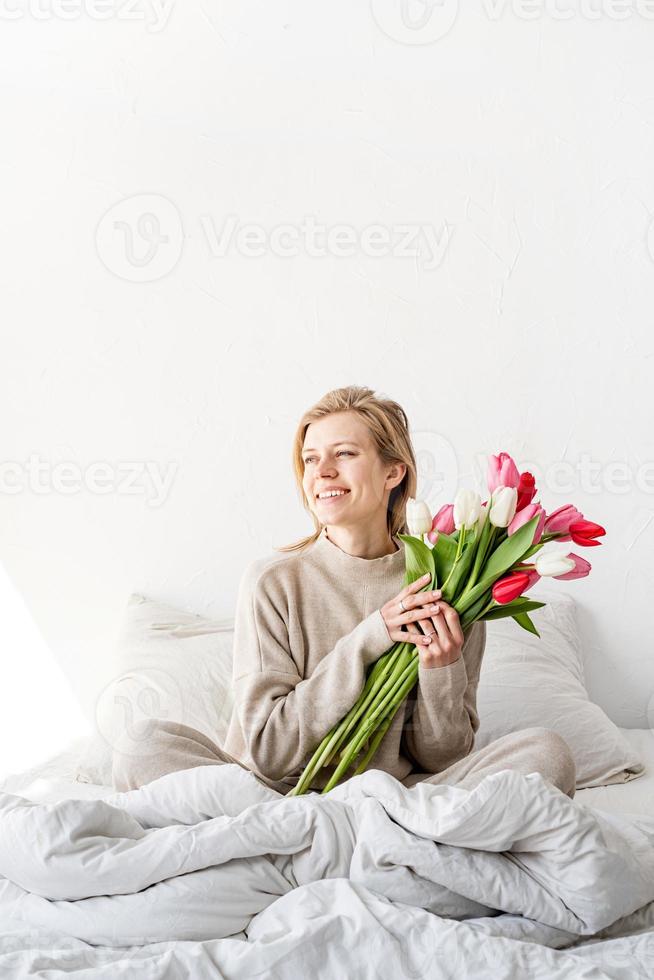 mulher sentada na cama de pijama segurando um buquê de flores de tulipa foto