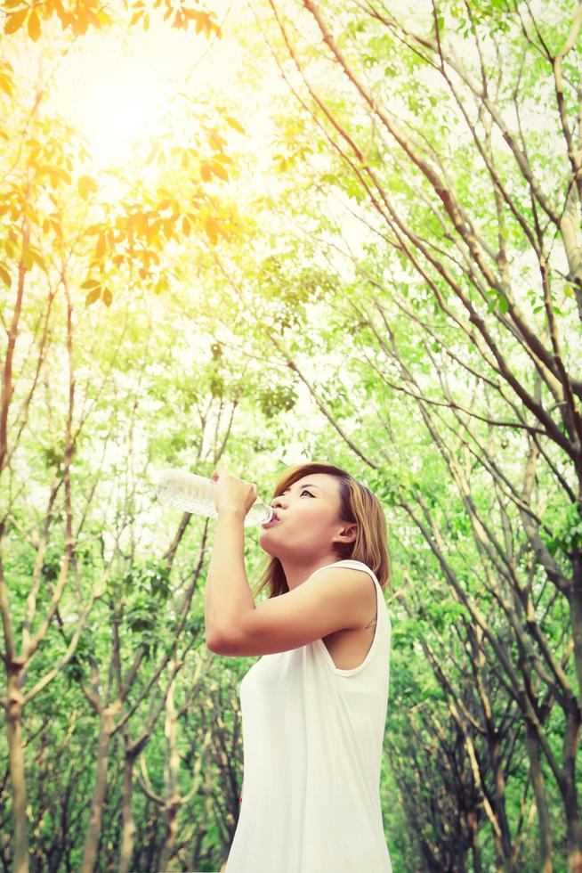 jovem mulher com vestido branco bebendo água na floresta verde. foto