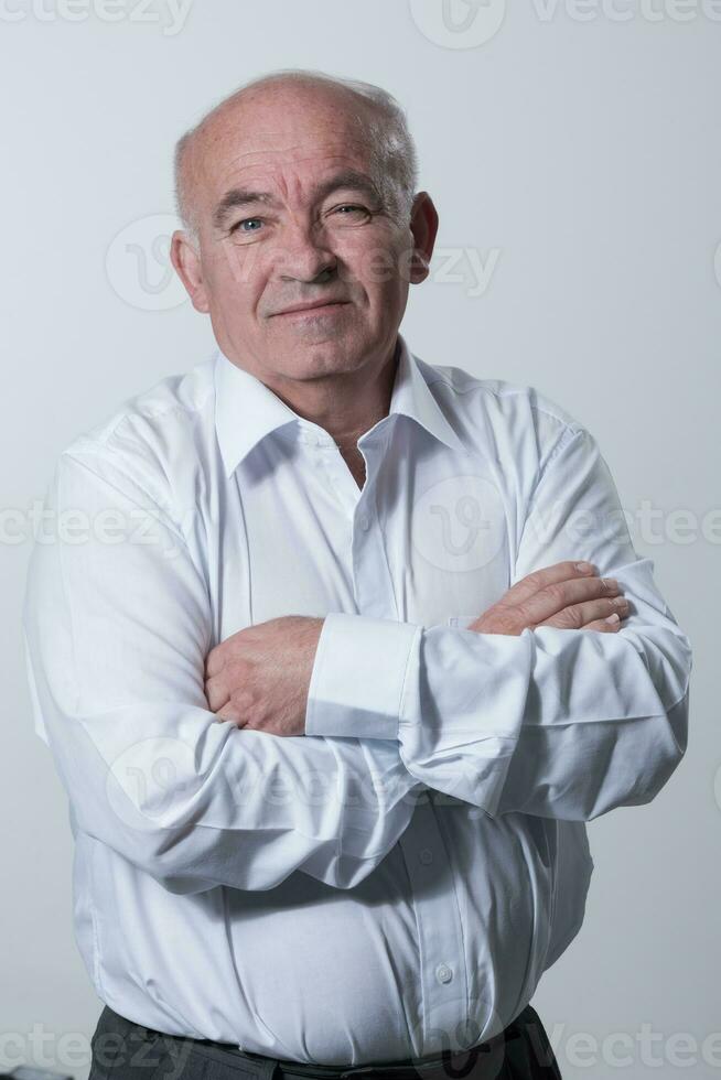 confiante Senior homem dentro branco camisa cruzando mãos em peito e olhando às Câmera enquanto em pé contra cinzento fundo. auto confiante Senior isolado branco estúdio atirar. foto