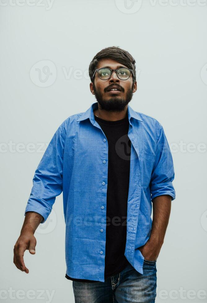indiano sorridente jovem homem com azul camisa e óculos posando em cinzento fundo foto
