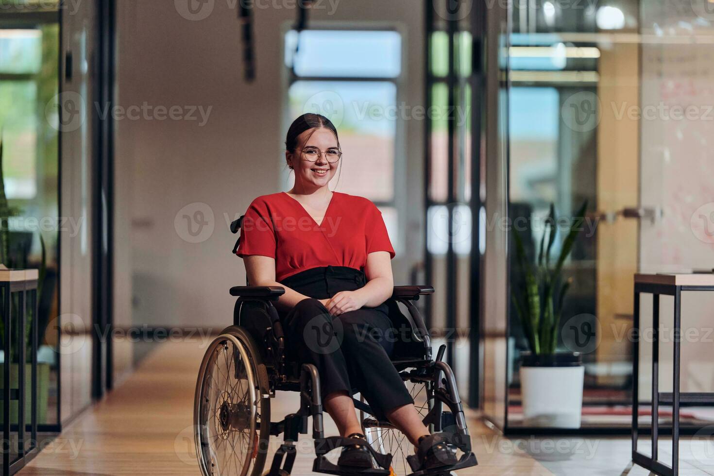 uma moderno jovem empresária dentro uma cadeira de rodas é cercado de a inclusivo área de trabalho com com paredes de vidro escritórios, incorporando determinação e inovação dentro a o negócio mundo foto