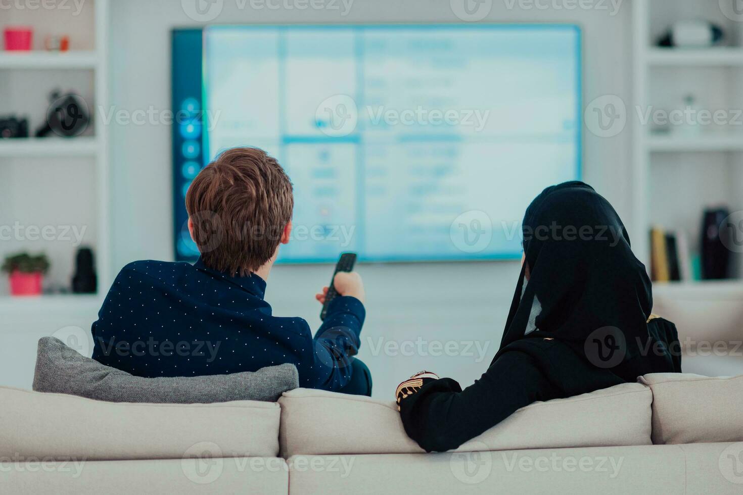 jovem muçulmano casal mulher vestindo islâmico hijab roupas sentado em sofá assistindo televisão juntos durante a mês do Ramadã às moderno casa foto