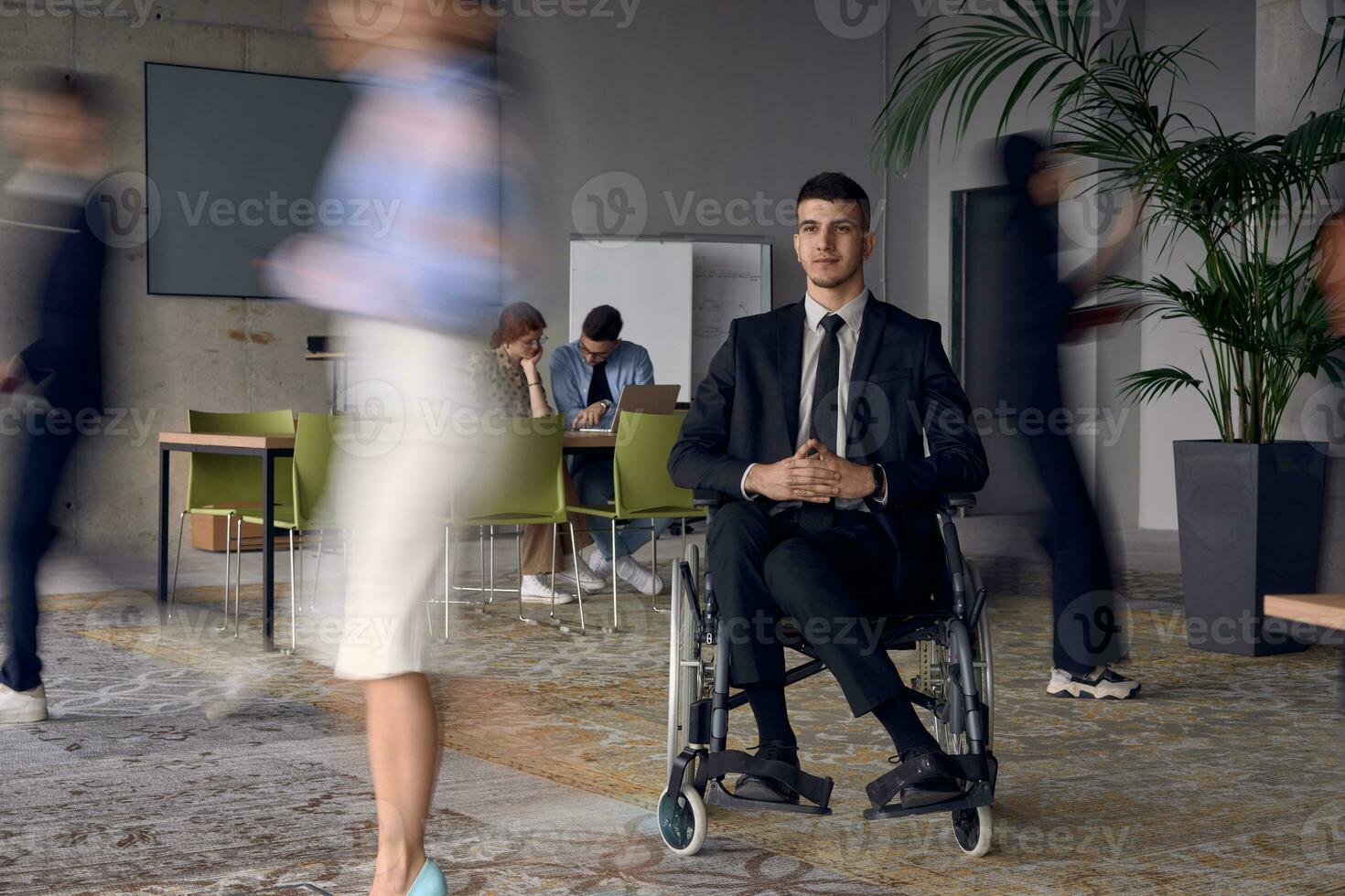 uma homem de negocios dentro uma cadeira de rodas dentro uma moderno escritório, cercado de dele colegas quem estão retratado com borrado movimentos, simbolizando seus Apoio, suporte e solidariedade Como elas navegar a área de trabalho junto. foto